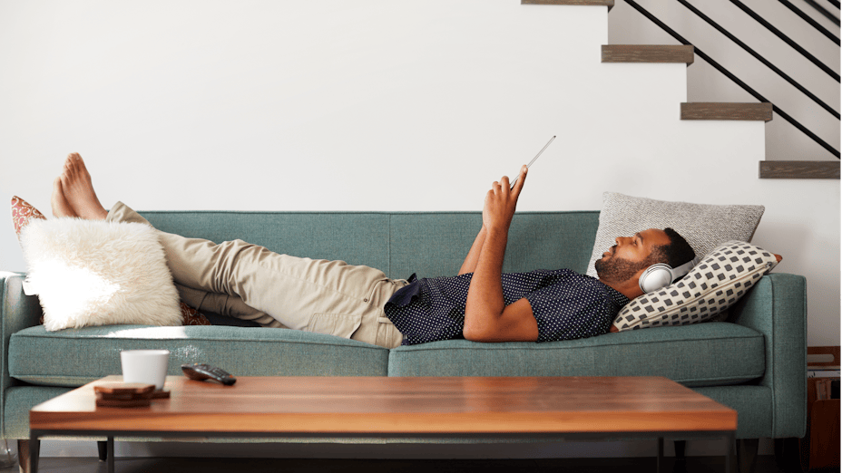 Man Lying On Sofa At Home Wearing Headphones to protect against noise.