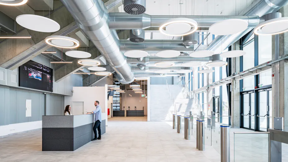 Modern public concourse with suspended circular acoustic rafts, exposed ventilation services, reception desk, ticket gates, and floor-to-ceiling windows bringing natural daylight into the space.