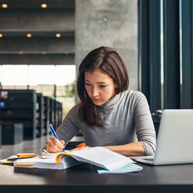 Female student taking notes from a book at library. Young asian woman sitting at table doing assignments in college library. People, school, acoustics.