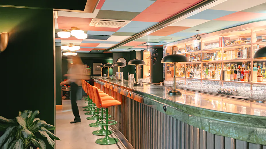 Bar interior with a colourful checkerboard acoustic ceiling, long green marble bar, orange bar stools, pendant lighting, and illuminated shelves stocked with bottles behind the counter.