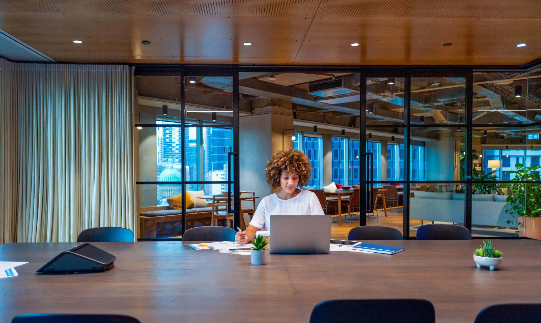 Young woman working on a laptop computer in a modern office. She is focused and holding a pen. There is paperwork and a phone on her desk. Stock photo ID:1590619701