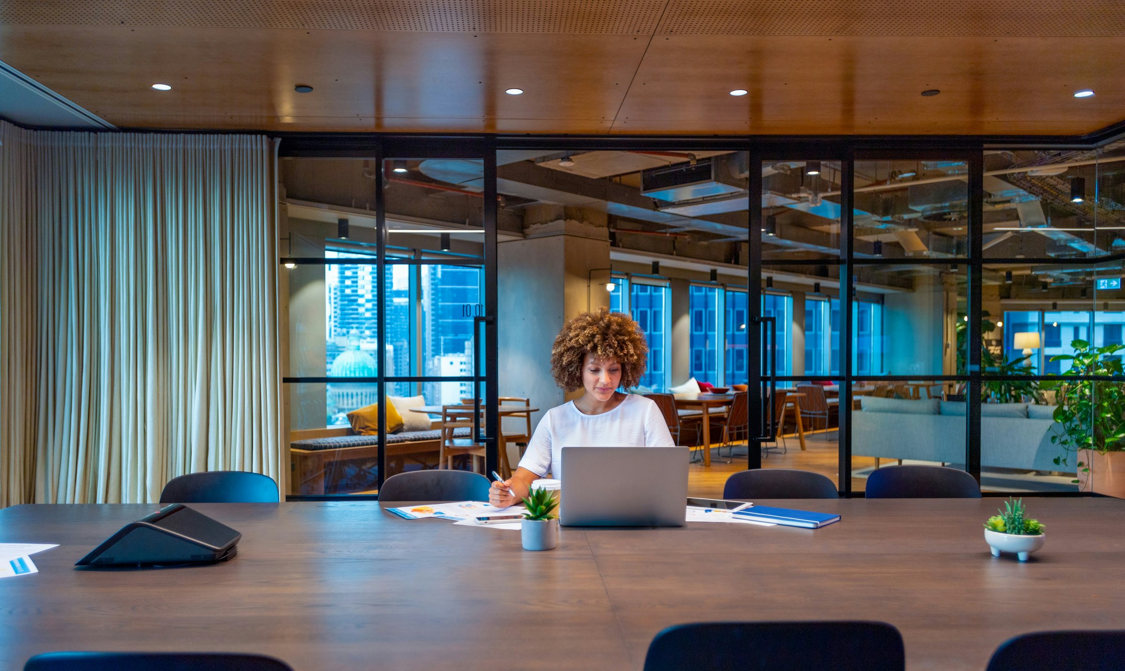 Young woman working on a laptop computer in a modern office. She is focused and holding a pen. There is paperwork and a phone on her desk. Stock photo ID:1590619701