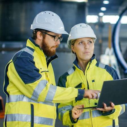Male and Female Industrial Engineers in Hard Hats Discuss New Project while Using Laptop. They Make Showing Gestures. They Work in a Heavy Industry Manufacturing Factory.
