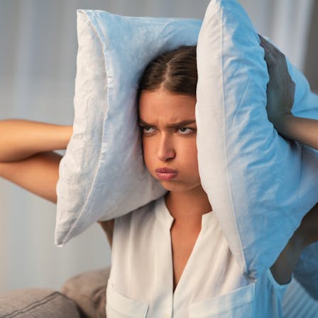 Annoyed Young Lady Covering Ears With Pillow Irritated Because Of Noise At Home.