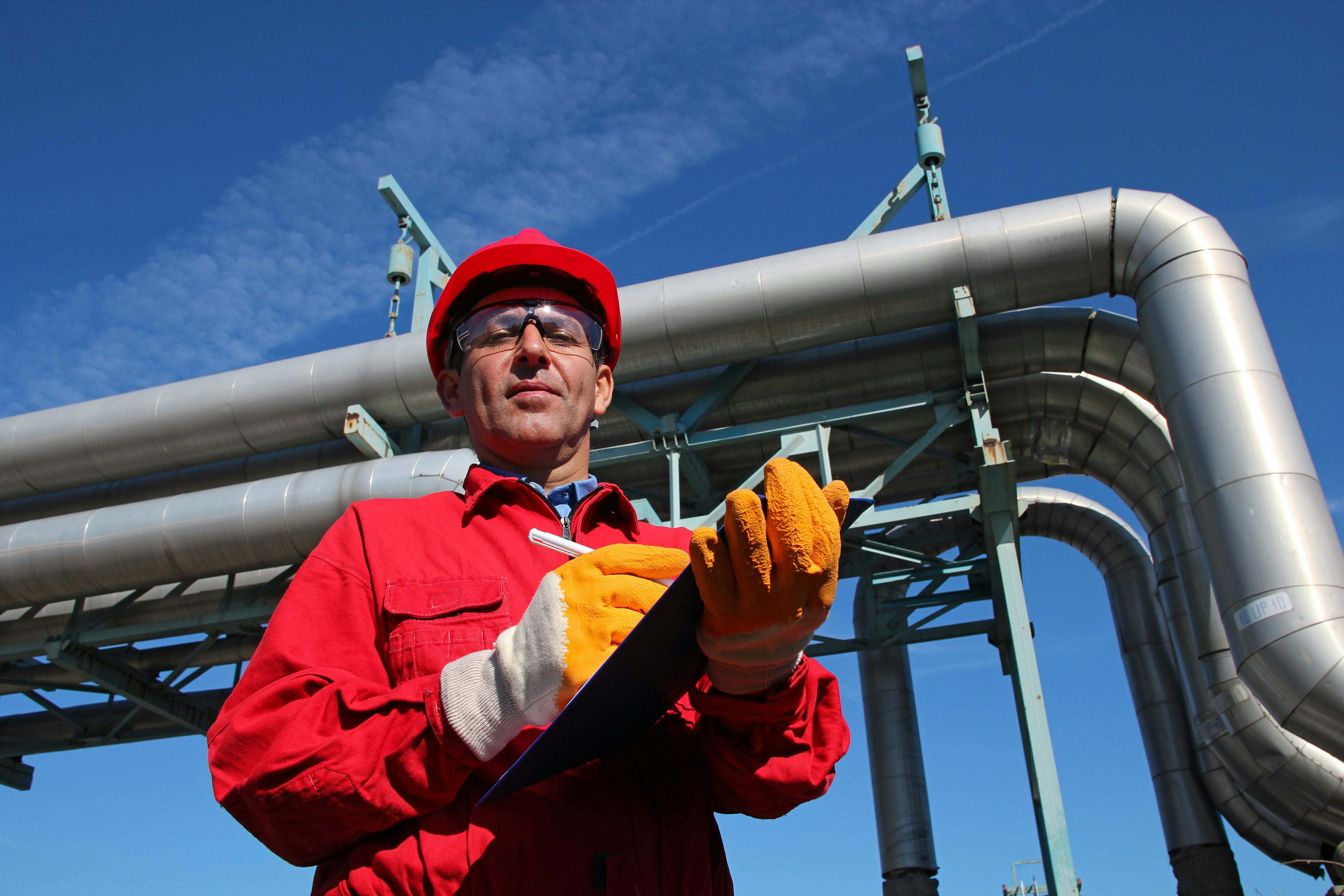 Worker wearing red overalls and hardhat , writing on clipboard next to pipelines.