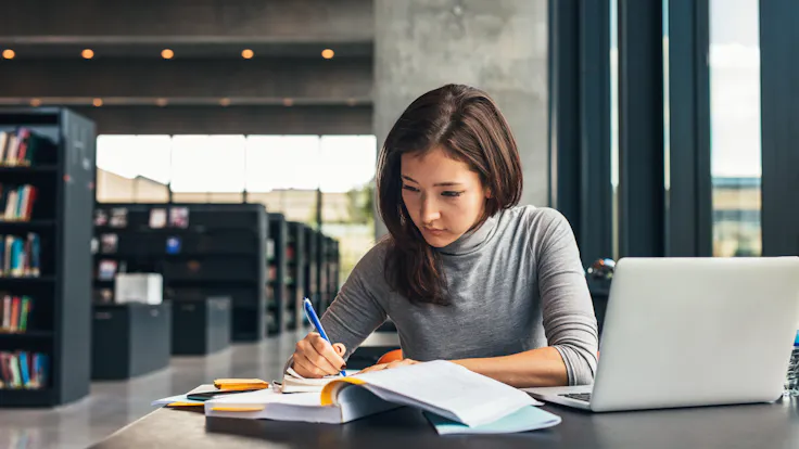 Female student taking notes from a book at library. Young asian woman sitting at table doing assignments in college library. People, school, acoustics.